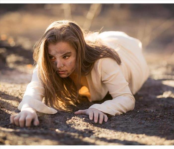 Young mixed race woman in white dress crawls on ground by trees destroyed by wildfire while covered in ashes